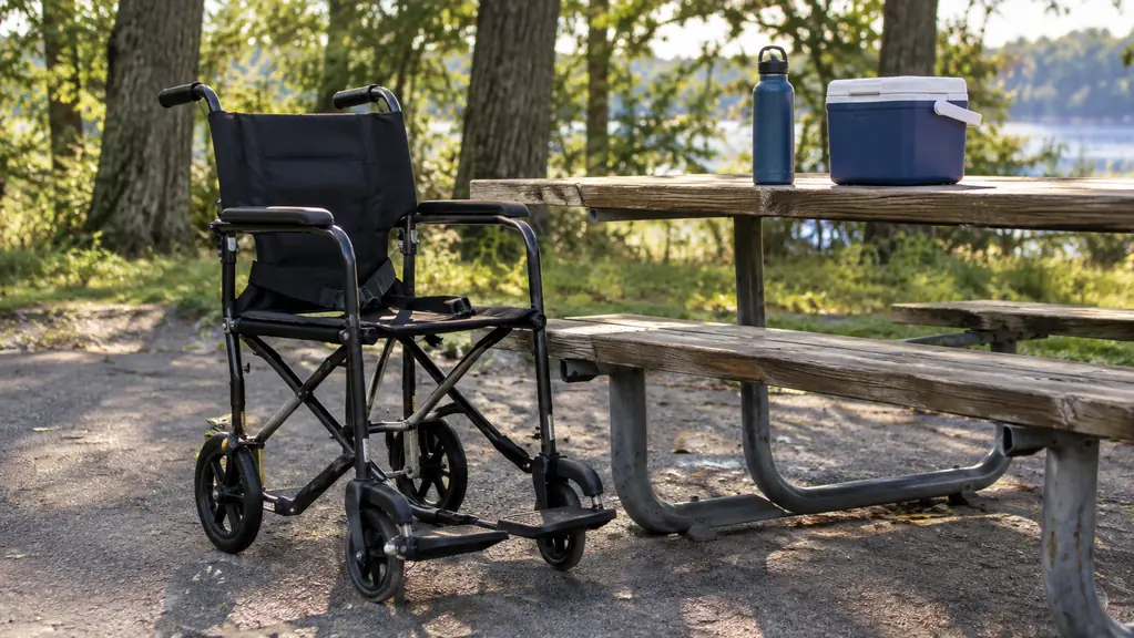 A transport chair parked beside a picnic table near a paved path, with a water bottle and a small cooler on the table