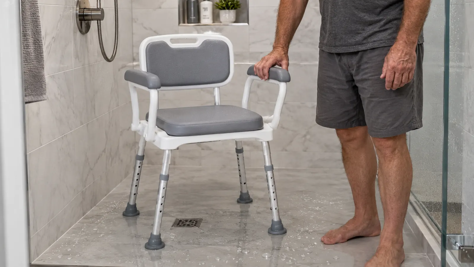 A shower chair with padded armrests and a back placed inside a walk-in shower, with a hand resting on the armrest